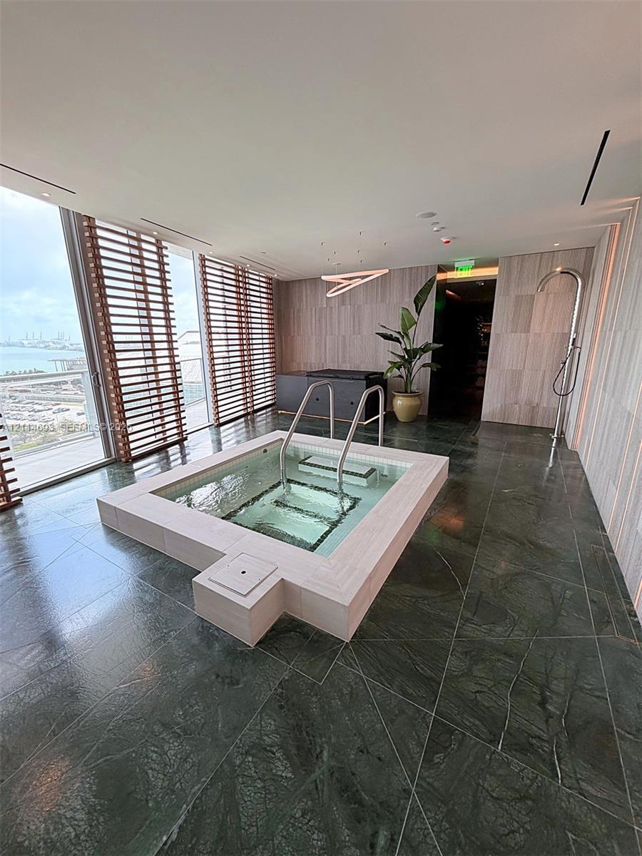 Built-in soaking tub with metal handrails, dark stone tile flooring, light wood-paneled wall accents, and floor-to-ceiling windows with wood slat blinds