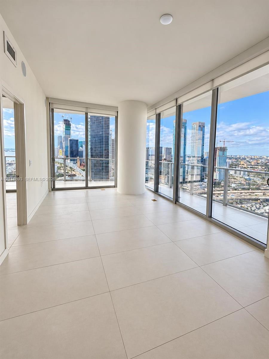 Floor-to-ceiling glass windows, neutral tile flooring, structural pillar, and balcony access with expansive city and water skyline surroundings