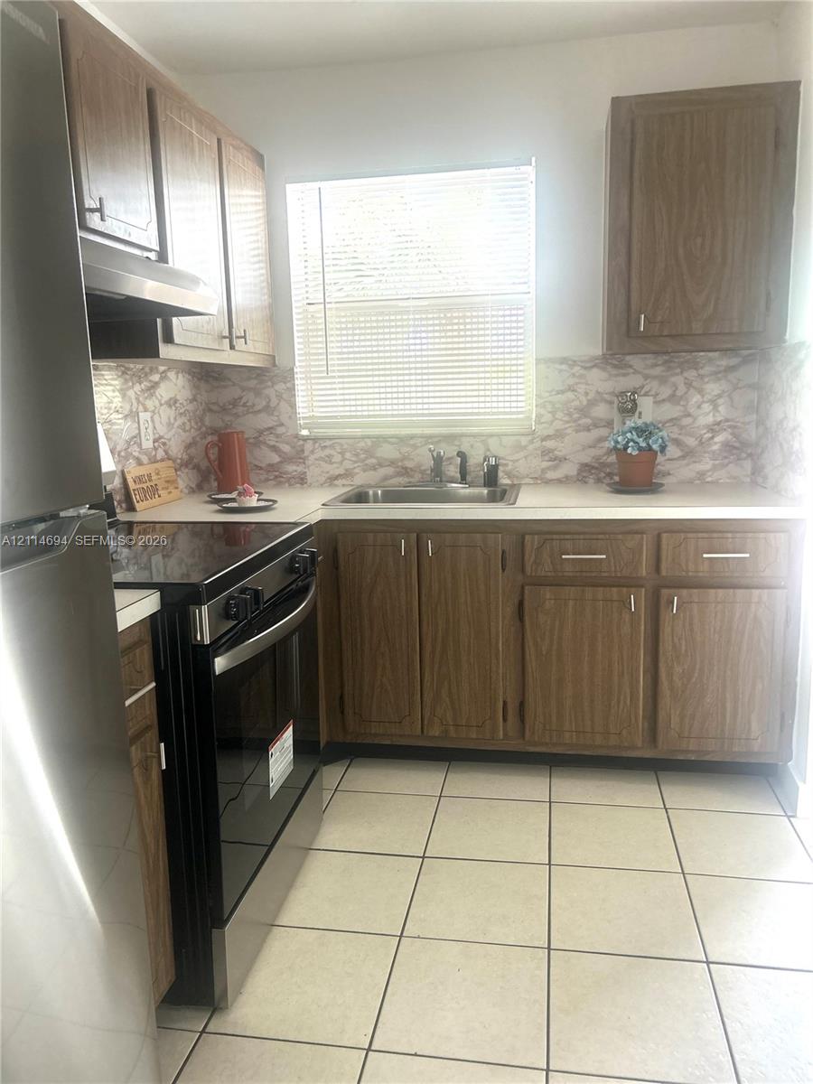 Kitchen featuring wood-finish cabinetry, stainless steel range and refrigerator, stone-patterned backsplash, and ceramic tile flooring