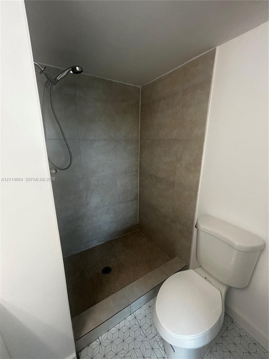 Shower stall featuring large-format neutral-toned tile walls and a handheld shower head, complemented by a floor with geometric patterned tile and a standard-profile toilet