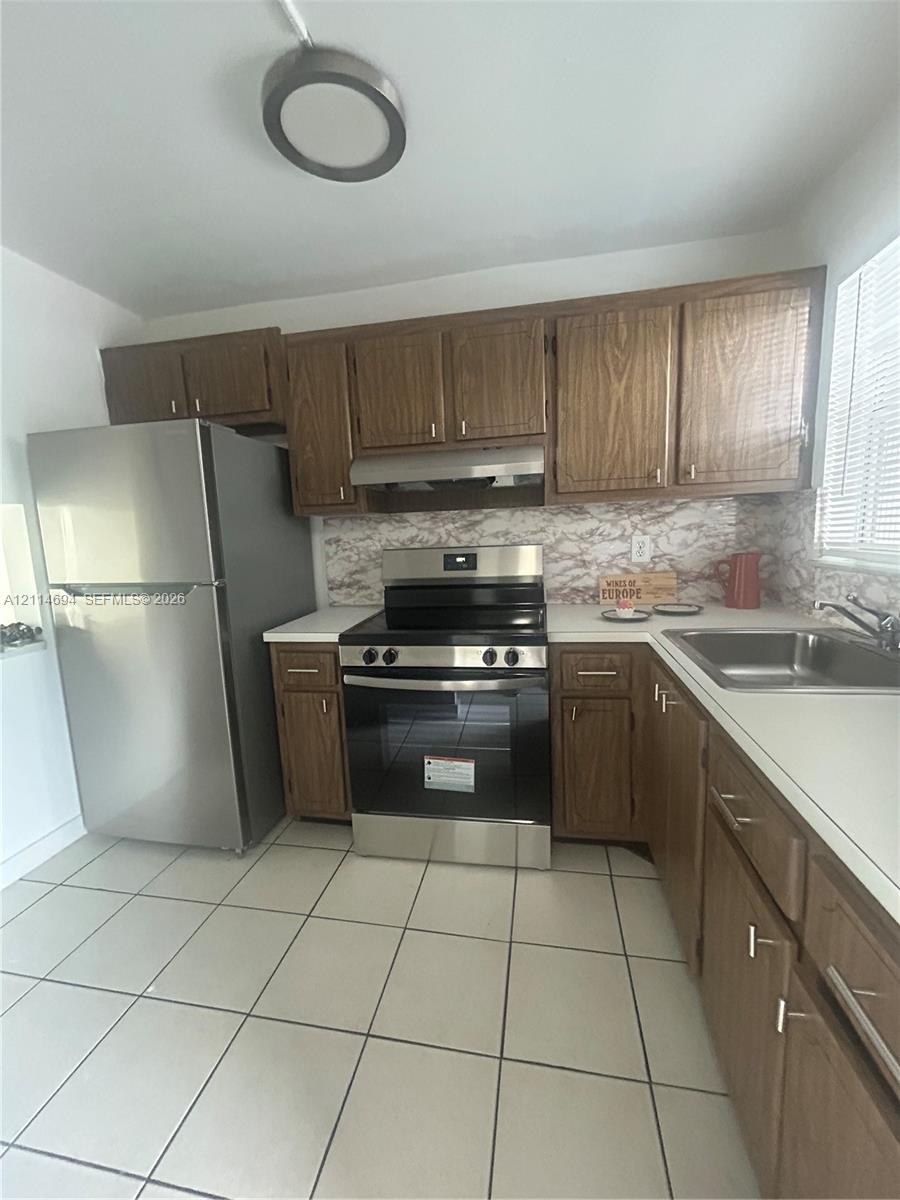 Kitchen featuring wood-finish cabinetry, stainless steel refrigerator and range, tiled flooring, and a stone-finish backsplash