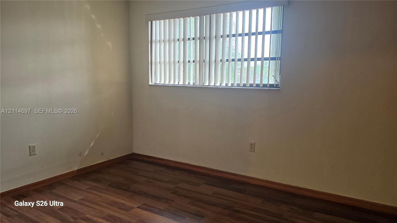 Interior space featuring neutral-toned walls, wood-finish flooring, and a large horizontal window equipped with vertical blinds