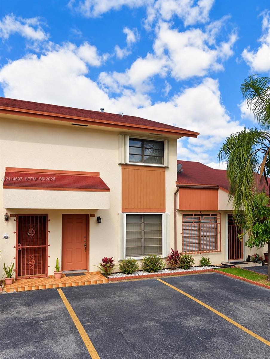 Two-story exterior featuring a light-toned stucco finish, red shingle roof, integrated metal security doors, tiled entry landing, and designated asphalt parking