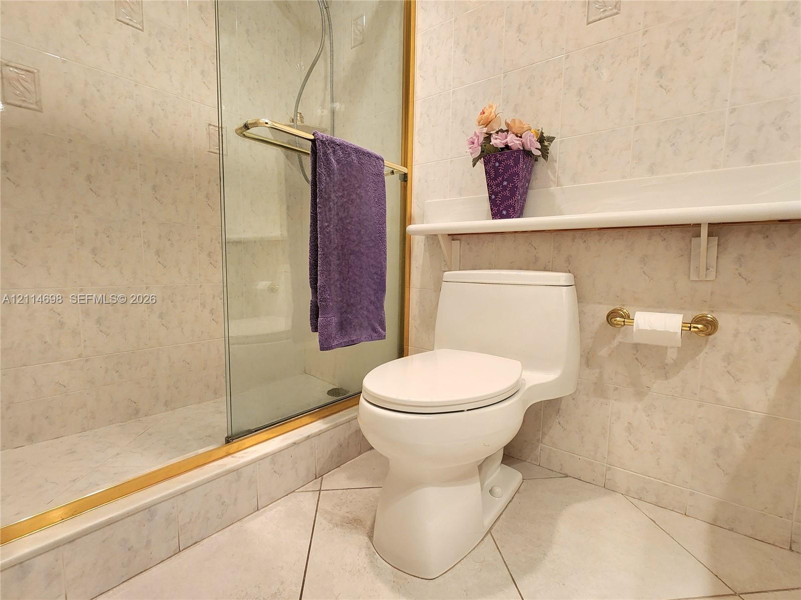 Glass-enclosed shower with gold-tone metal trim, wall-mounted built-in shelving, low-profile toilet, and floor-to-ceiling tile work