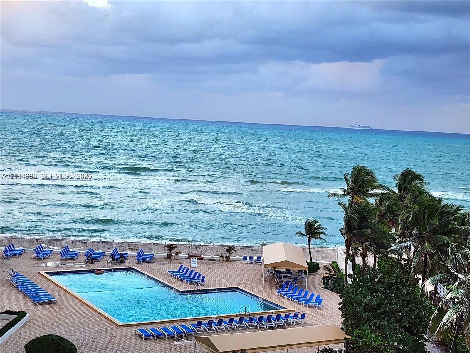 Outdoor swimming pool with a paved deck, permanent shaded structures, and mature palms overlooking the ocean