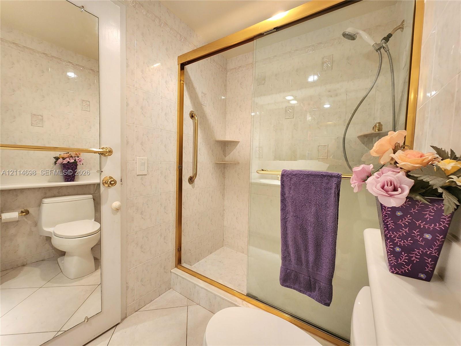 Walk-in shower featuring a glass enclosure with gold-tone hardware, tile-surround walls, and built-in shelving, complemented by a ceramic toilet and large wall-mounted mirror