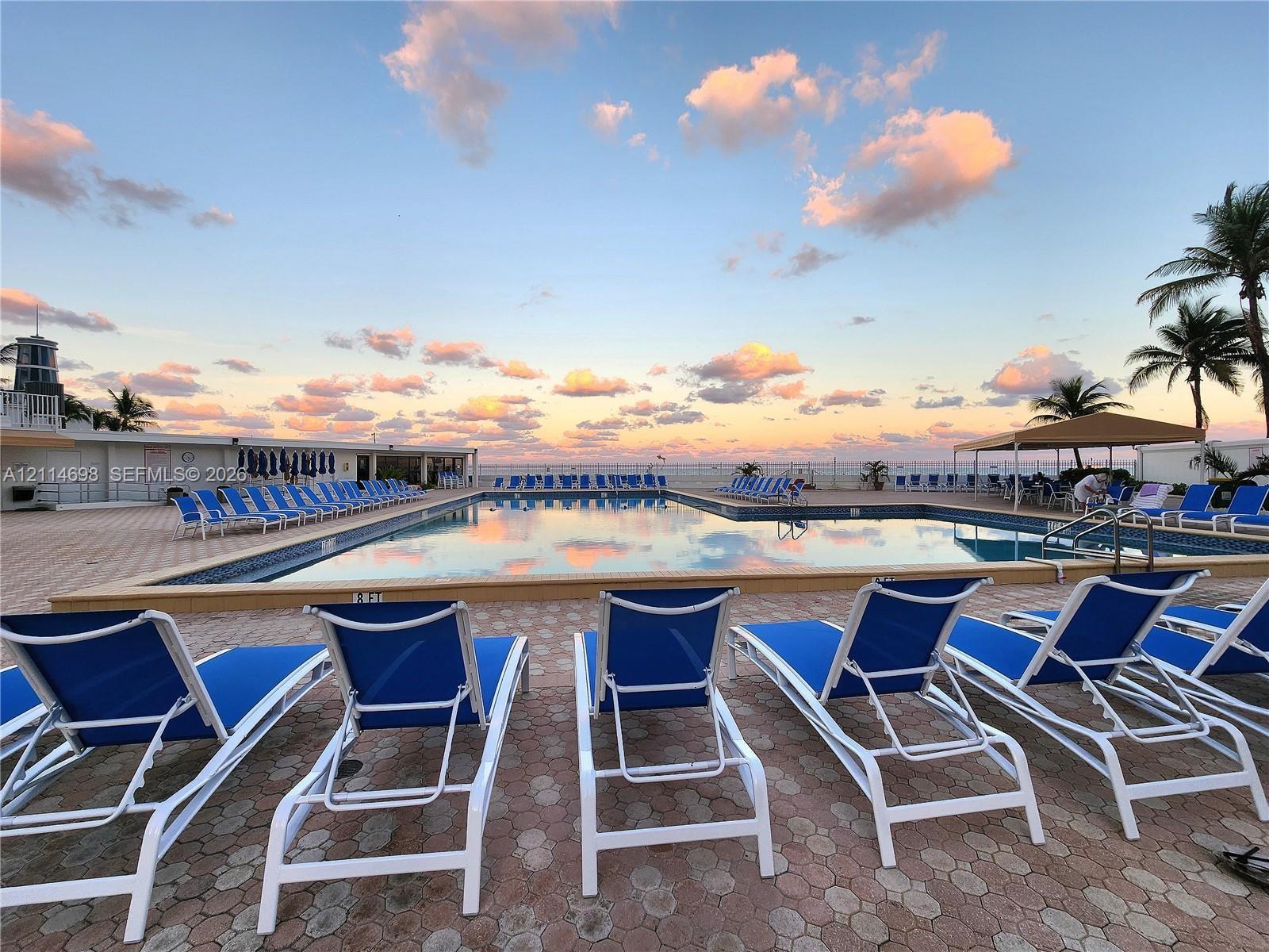 In-ground swimming pool featuring a geometric tiled deck, surrounding perimeter fencing, and a permanent shade structure