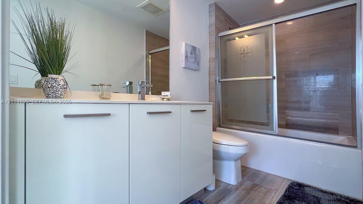 Modern vanity with white cabinetry and a solid surface countertop, featuring a single-lever faucet, large wall mirror, and a tub-shower combination with sliding glass doors and neutral-toned wall tiling