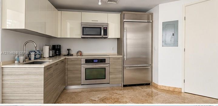 Kitchen featuring high-gloss upper cabinetry, wood-finish lower cabinetry, integrated stainless steel refrigerator, built-in wall oven with cooktop, and stone tile flooring