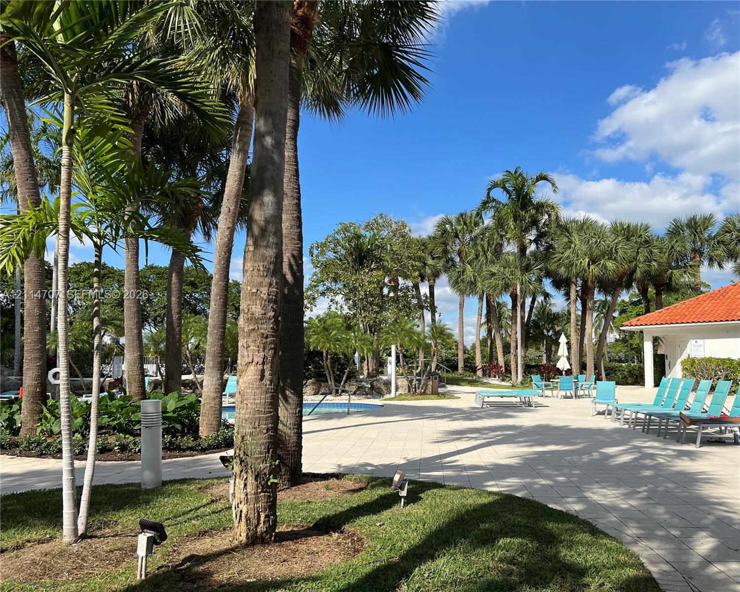 Outdoor terrace featuring light-toned paved hardscaping, mature palm trees, manicured landscaping, and a red-tiled roof structure