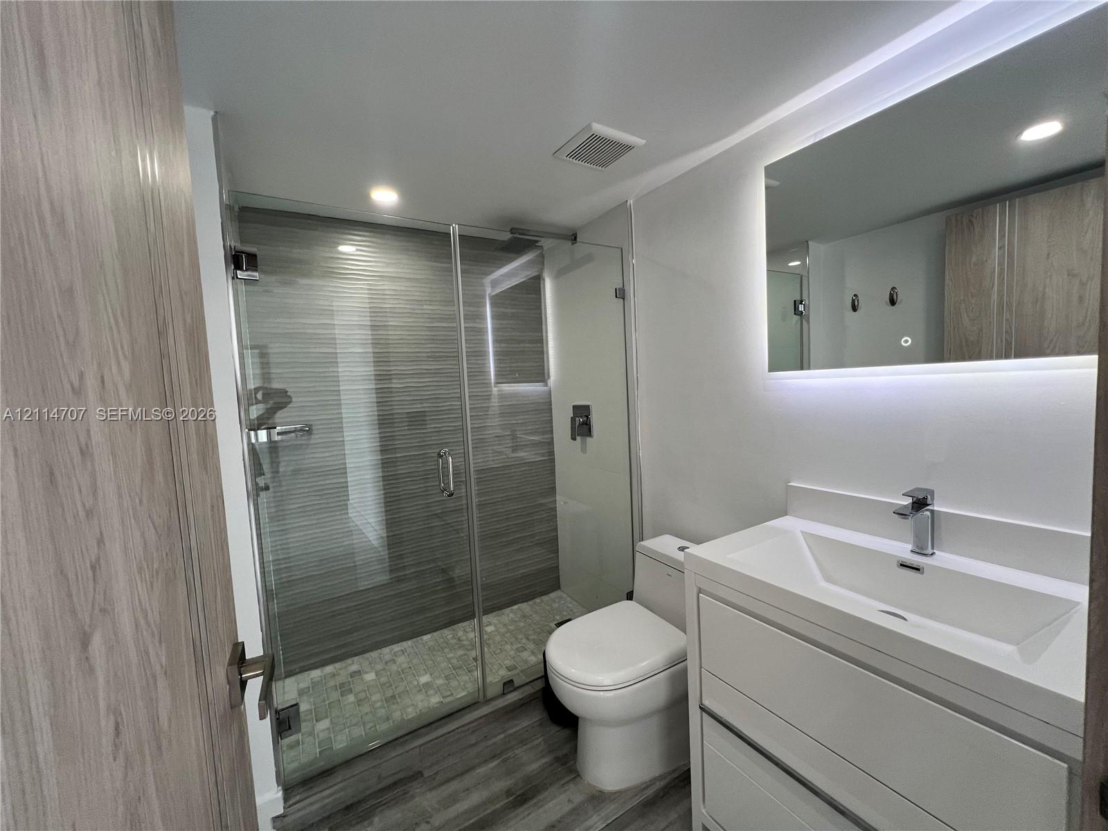Modern bathroom featuring a glass-enclosed walk-in shower with horizontal tile surround, a built-in vanity with a rectangular sink, wood-finish cabinetry, and an integrated LED-lit mirror