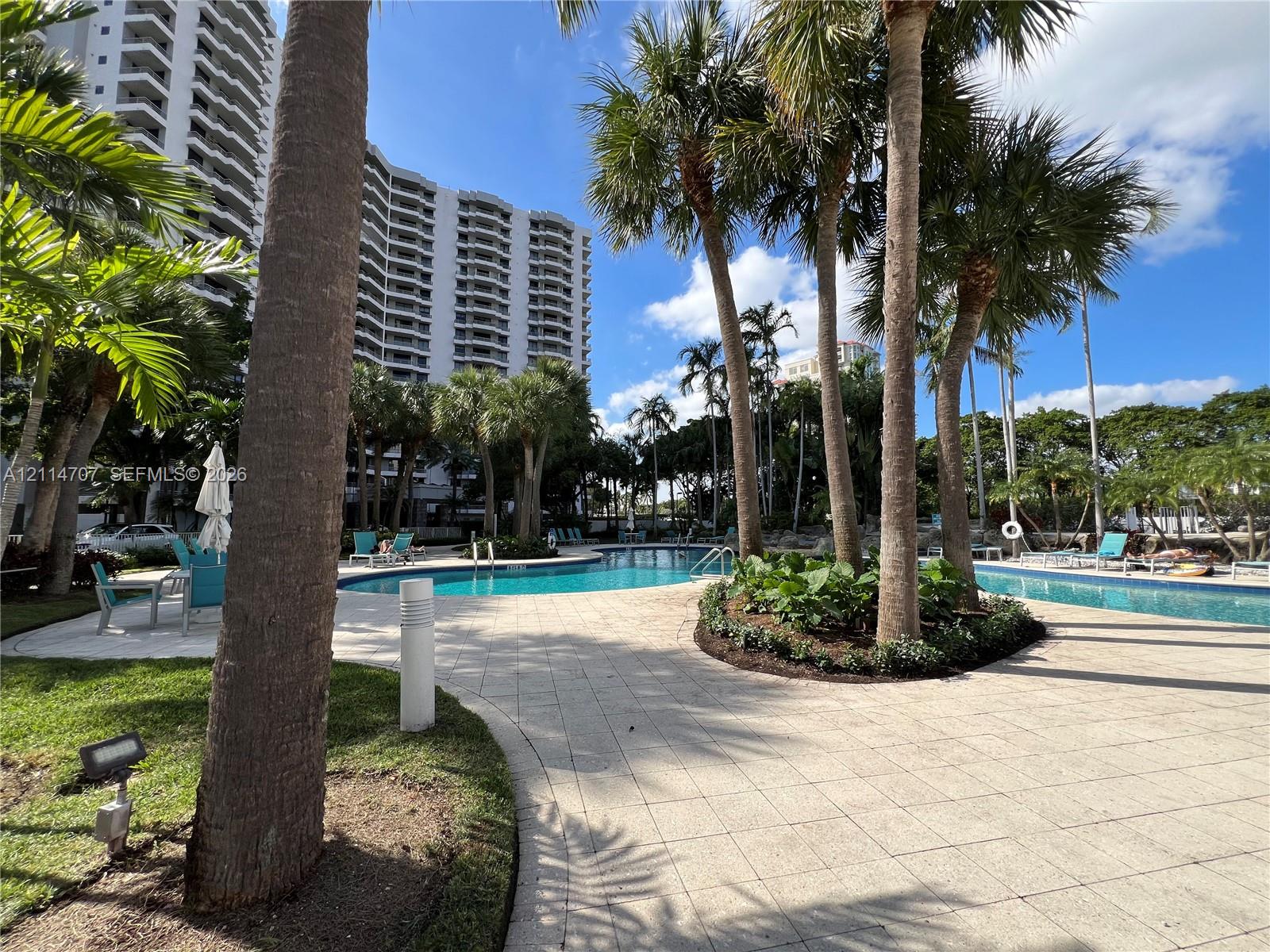 Outdoor swimming pool area featuring paved stone decking, mature palm trees, landscaped garden beds, and metal ladder access