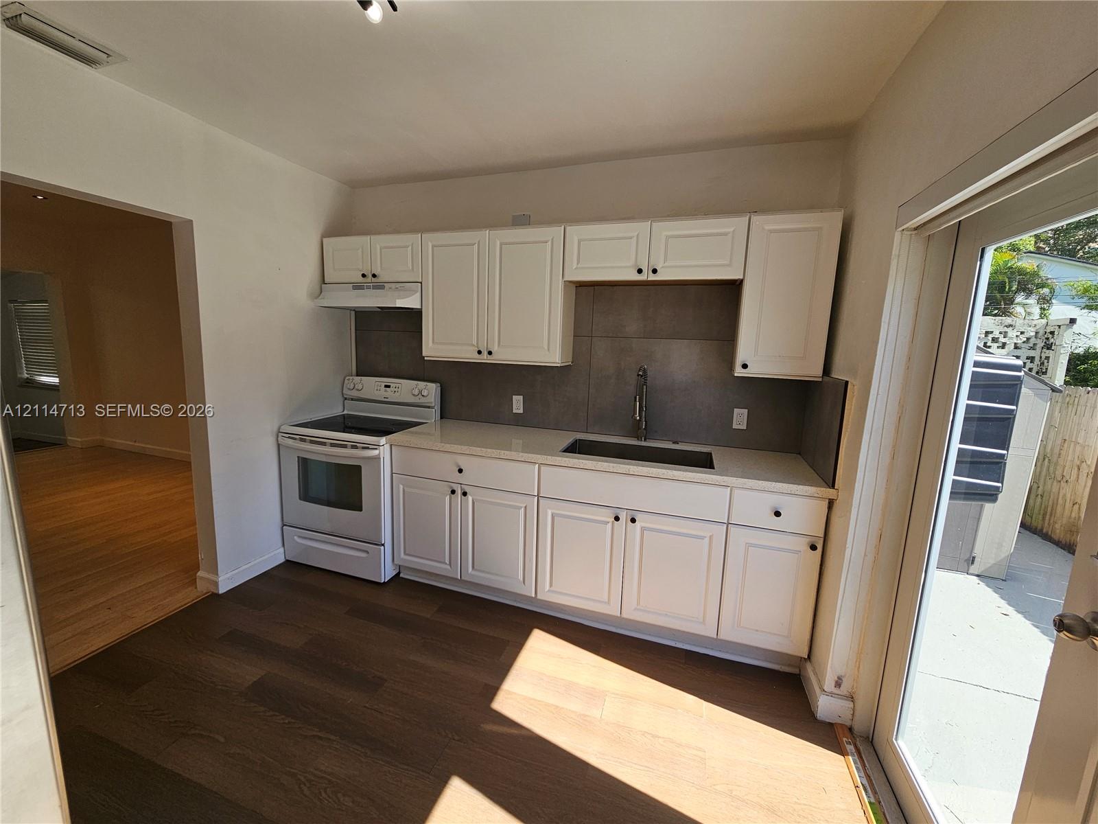 Kitchen featuring white shaker-style cabinetry, neutral-toned solid surface countertops, a matte tile backsplash, a stainless steel sink with a high-arch faucet, and dark wood-finish flooring