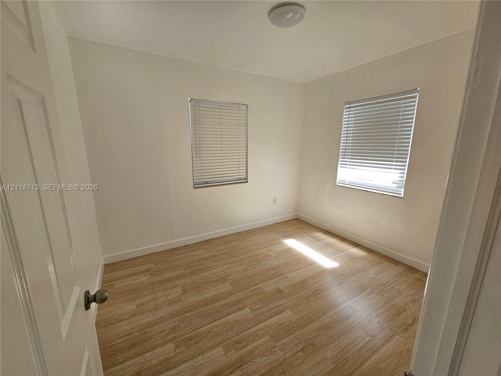 Neutral interior room featuring wood-finish flooring, two windows with horizontal blinds, and a ceiling-mounted light fixture