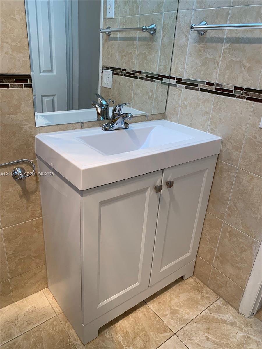 White vanity cabinet with a rectangular basin, metallic hardware, wall-mounted mirror, and tile wall surfacing featuring a decorative mosaic border