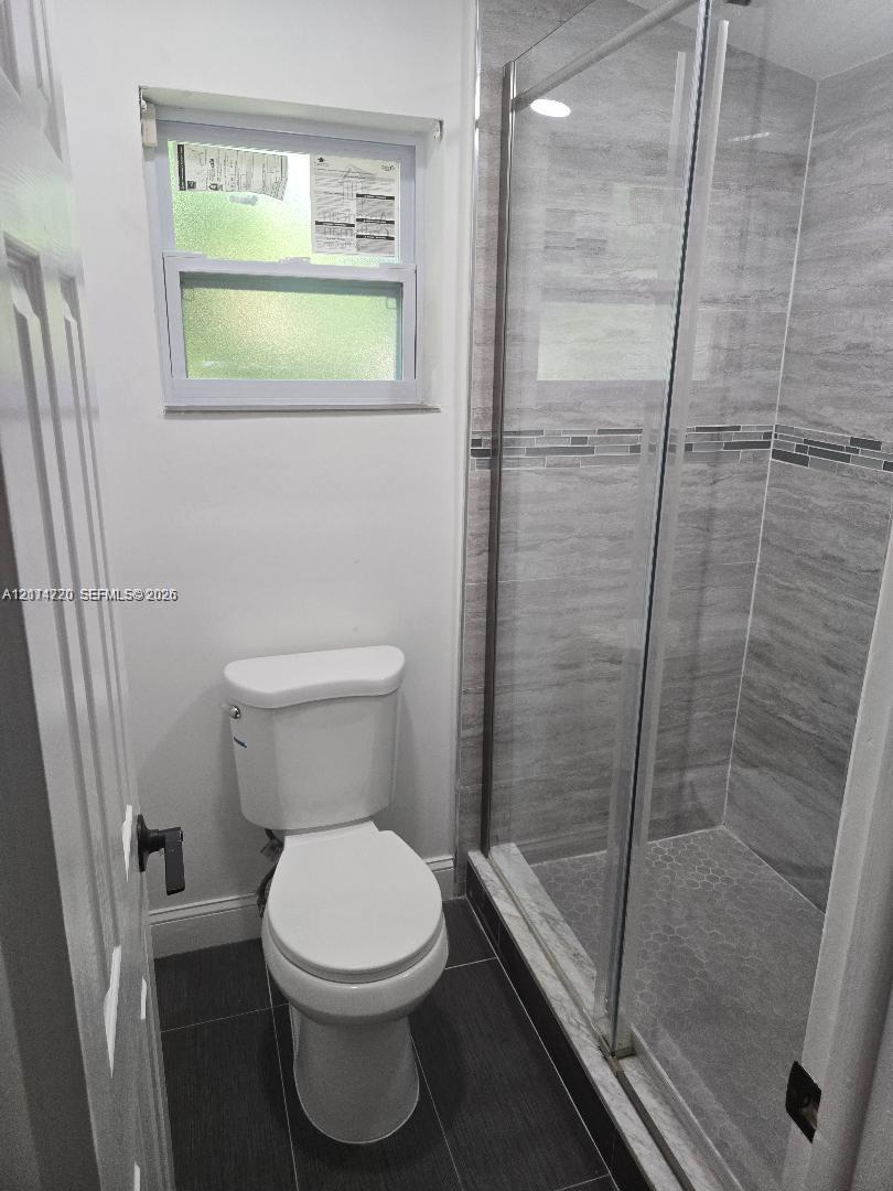 Glass-enclosed shower with horizontal wall tile, mosaic accent strip, and dark floor tiling, complemented by a single-flush toilet and a horizontal sliding window