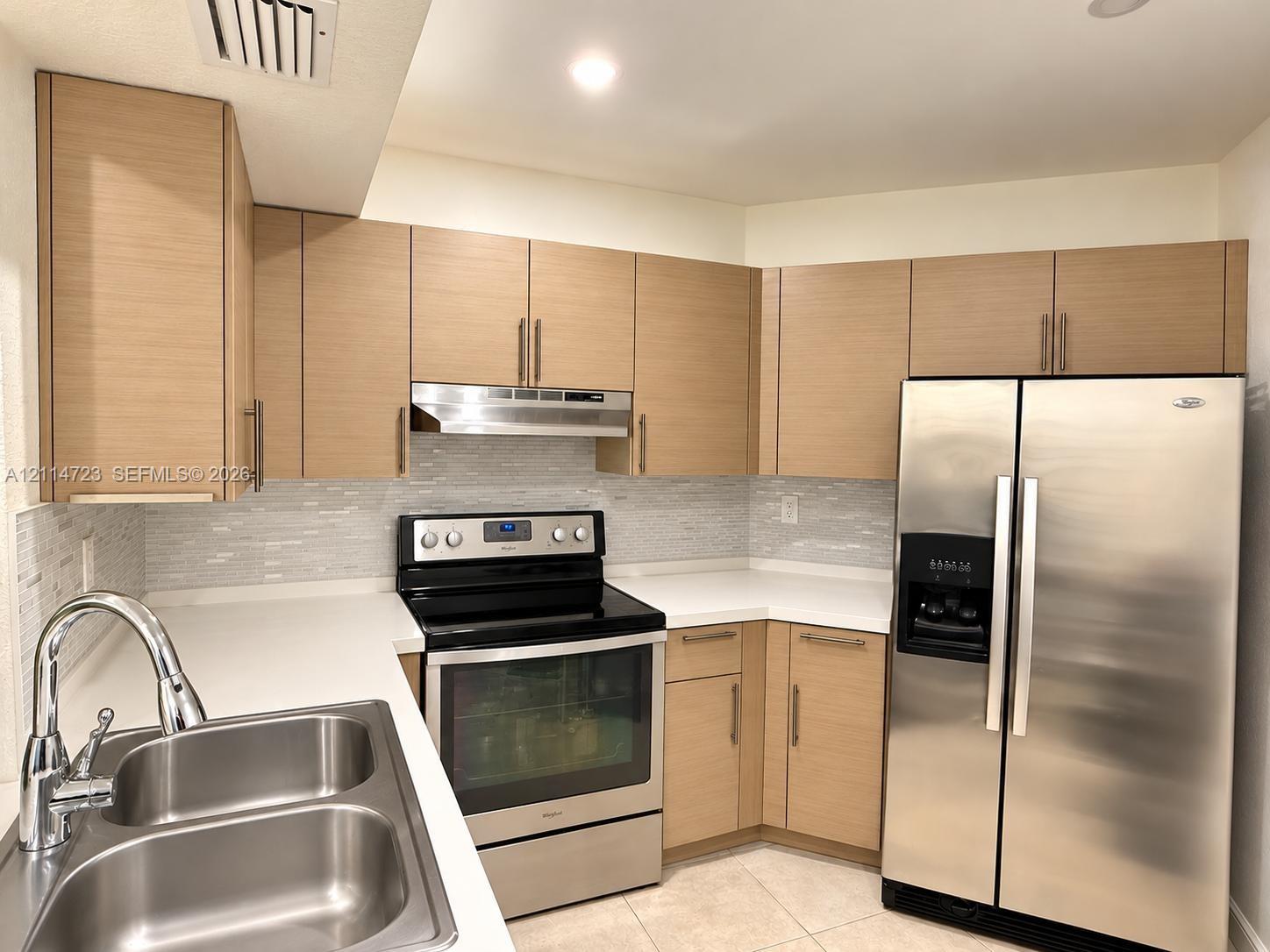 Kitchen featuring flat-panel wood-finish cabinetry, solid surface countertops, a mosaic tile backsplash, stainless steel appliances, and a dual-basin stainless steel sink with a gooseneck faucet Kitchen featuring flat-panel wood-finish cabinetry, solid surface countertops, a mosaic tile backsplash, stainless steel appliances, and a dual-basin stainless steel sink with a gooseneck faucet