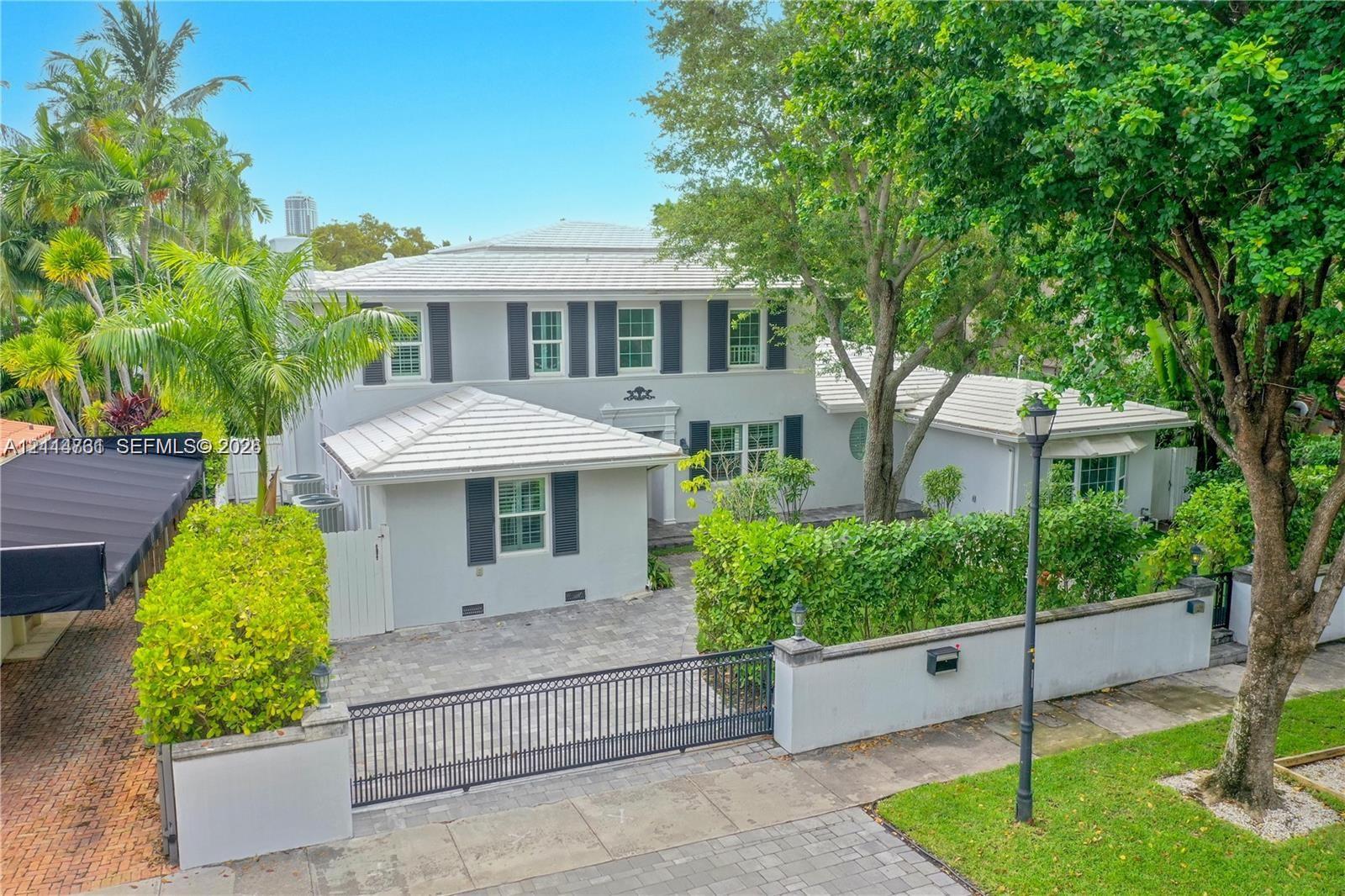 Two-story exterior featuring a light-toned stucco faade, metal roof, shuttered windows, paver driveway, and a gated perimeter wall