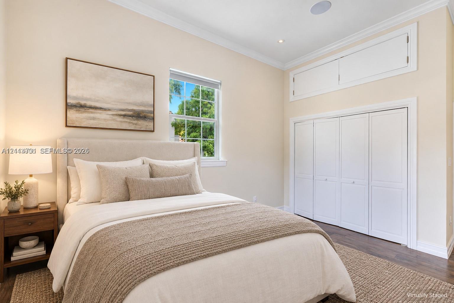 Neutral-toned room featuring dark wood-finish flooring, white bi-fold closet doors, additional overhead storage cabinetry, and a window with exterior greenery