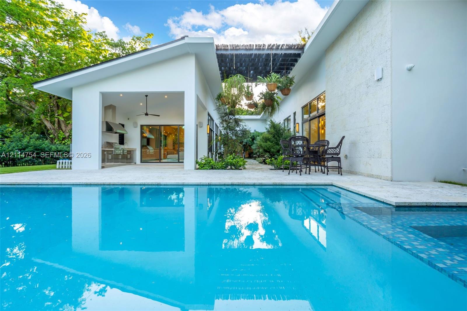 In-ground swimming pool with tiled border, expansive stone-paved patio, covered outdoor kitchen with stainless steel appliances, and minimalist white exterior siding