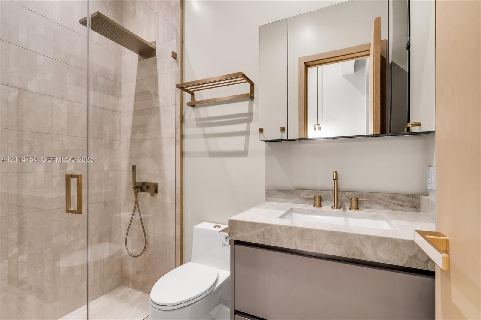 Modern vanity with a natural stone countertop and brass-finish faucet, featuring a mirrored medicine cabinet, a wall-mounted brass towel rack, and a glass-enclosed shower with decorative hexagonal tile wall covering and a rainfall showerhead