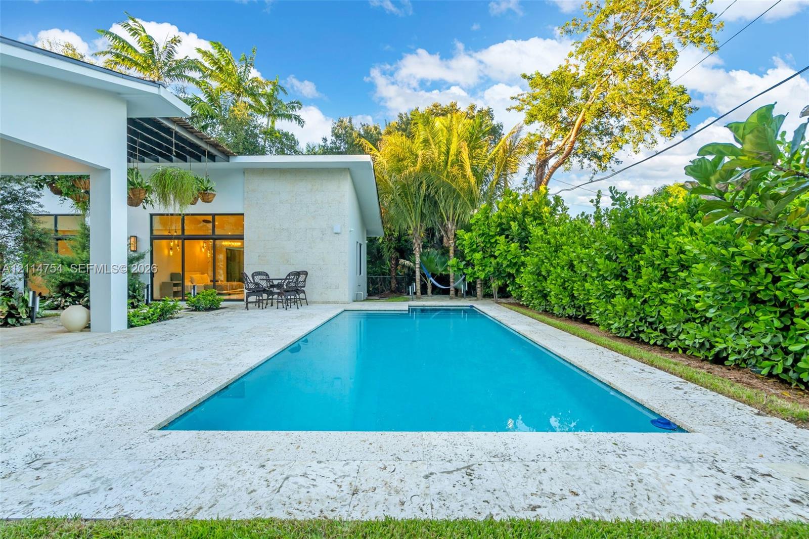 Covered patio featuring stone-tiled flooring, a built-in outdoor kitchen with stainless steel appliances, a slatted overhead architectural structure, and floor-to-ceiling glass sliding doors