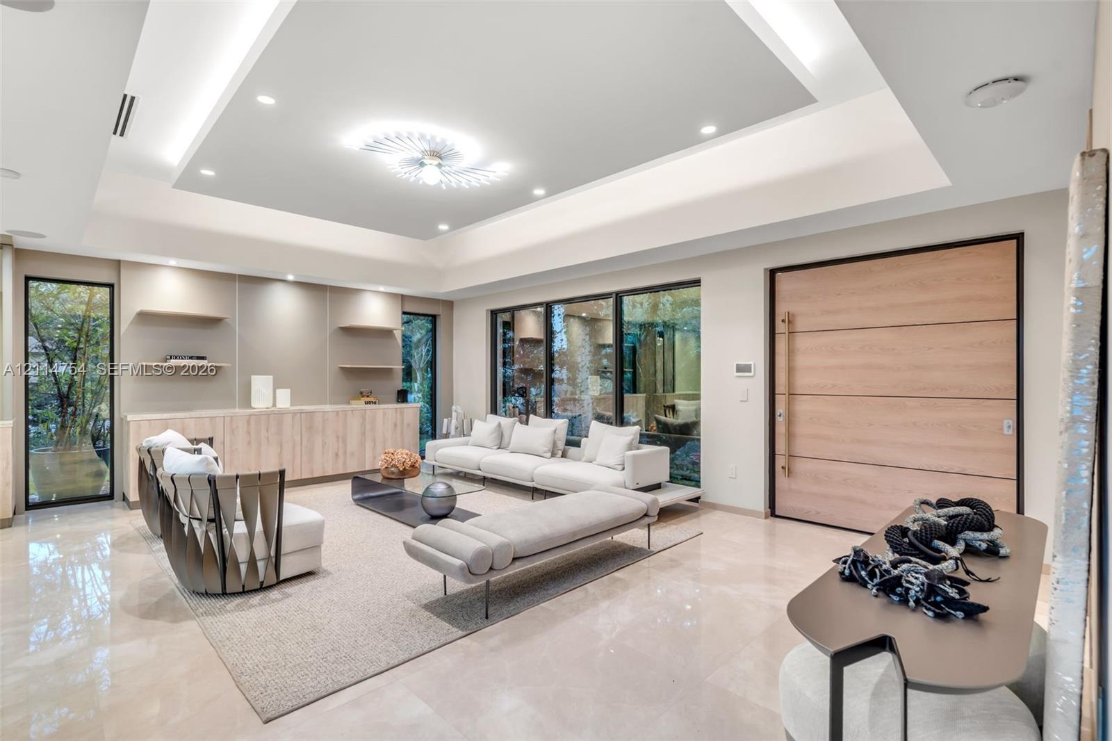 Expansive living area featuring a tray ceiling with recessed lighting, large-format polished flooring, floor-to-ceiling glass wall panels with dark frames, and distinctive modern tiered ceiling-mounted lighting