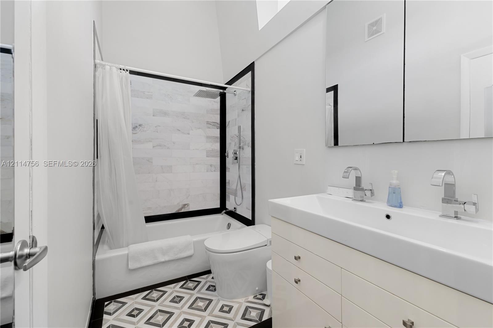 Bathroom featuring a white vanity with an integrated dual-faucet sink, tiled shower surround with black framing, geometric patterned floor tile, and a wall-mounted mirrored medicine cabinet