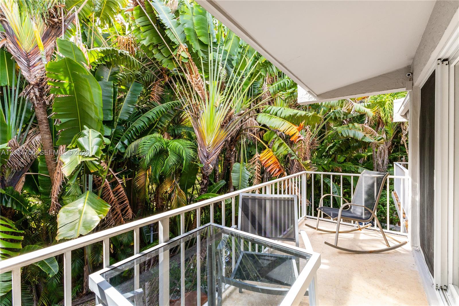 Covered balcony featuring white metal railings and concrete flooring, surrounded by lush tropical vegetation