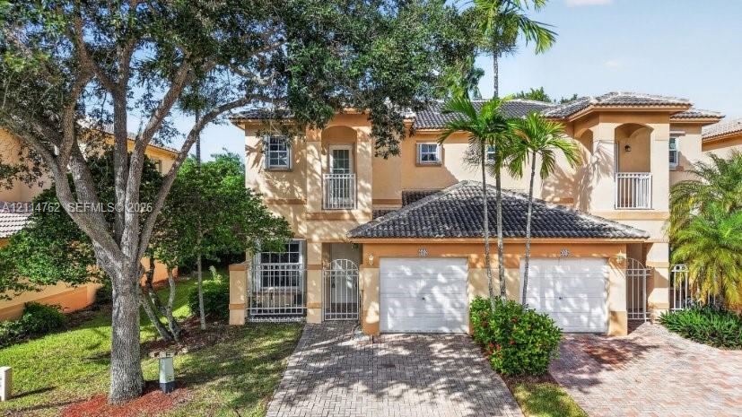 Stucco exterior featuring dual-level balconies with metal railings, a tiled roof, two-car garage bays, and a decorative brick paver driveway
