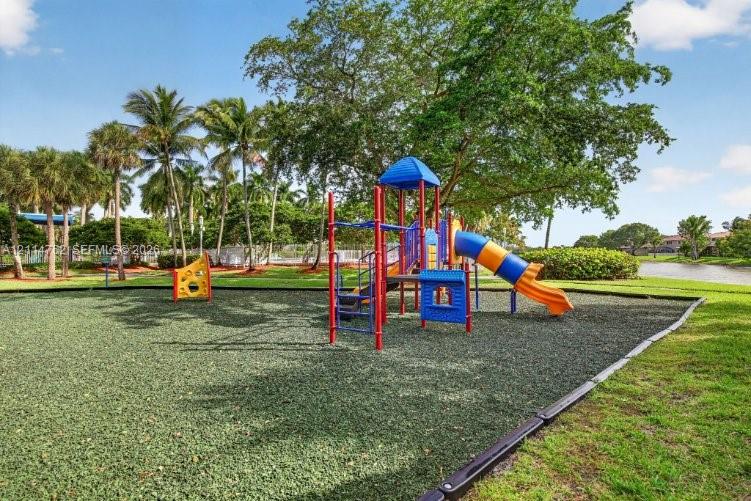 Outdoor recreation area featuring a multi-component play structure, rubberized ground surfacing, mature trees, and landscaping situated alongside a water feature