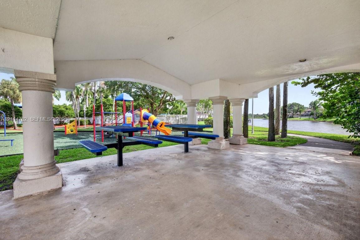 Covered pavilion with architectural columns, concrete flooring, and recreational space featuring playground equipment and paved walking paths