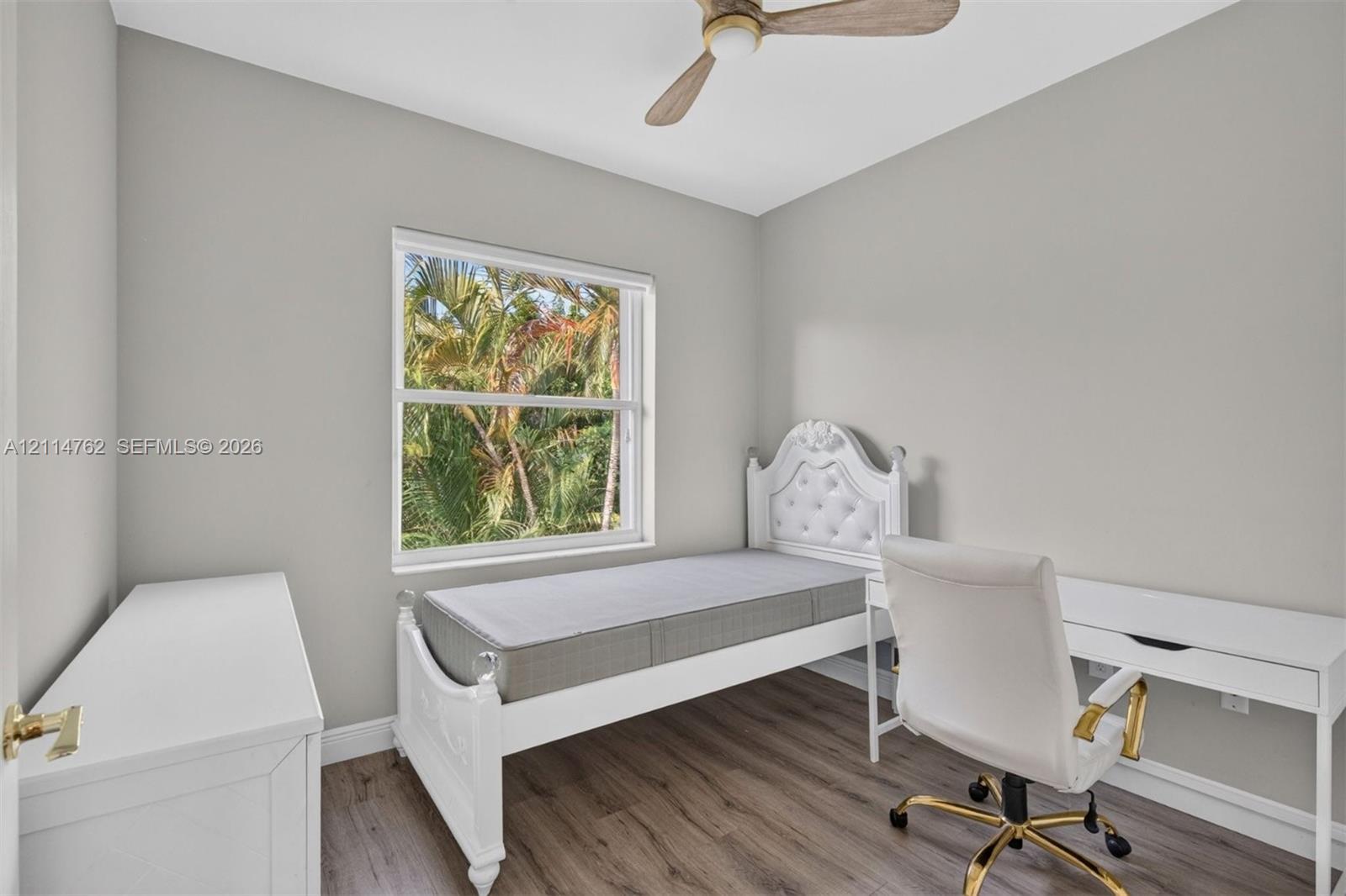 Neutral-toned interior space featuring wood-finish flooring, a ceiling fan with light fixture, and a large window providing natural light and exterior foliage