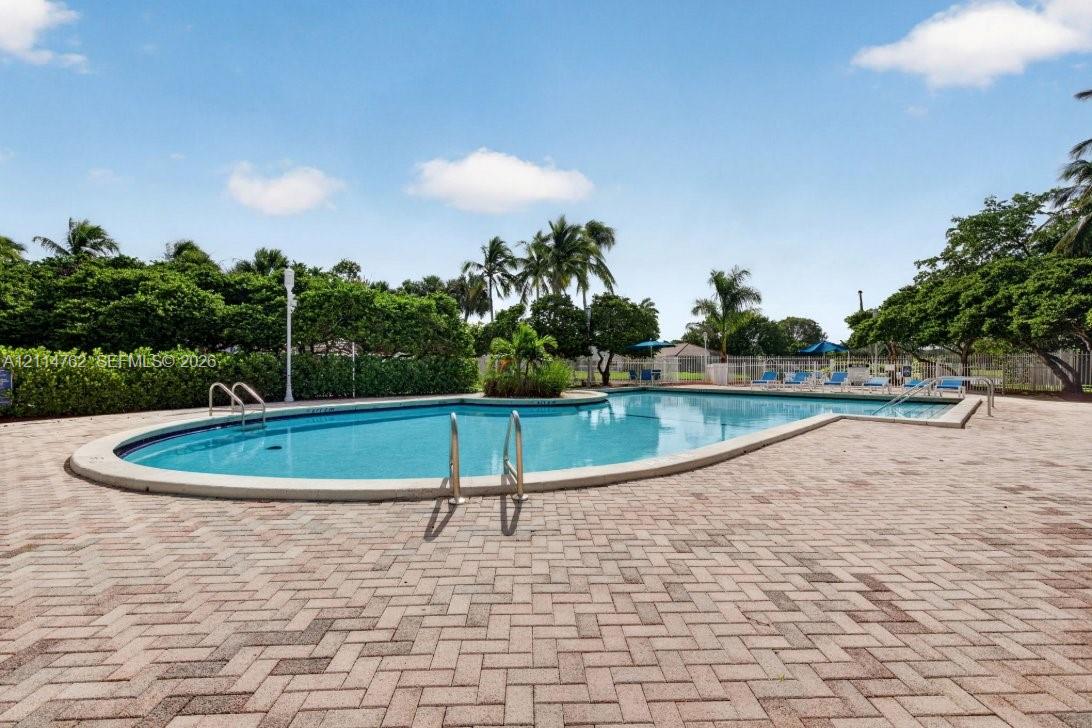 In-ground swimming pool featuring a curved design, metal handrails, and a surrounding herringbone-patterned paver deck bordered by established hedging and mature palms