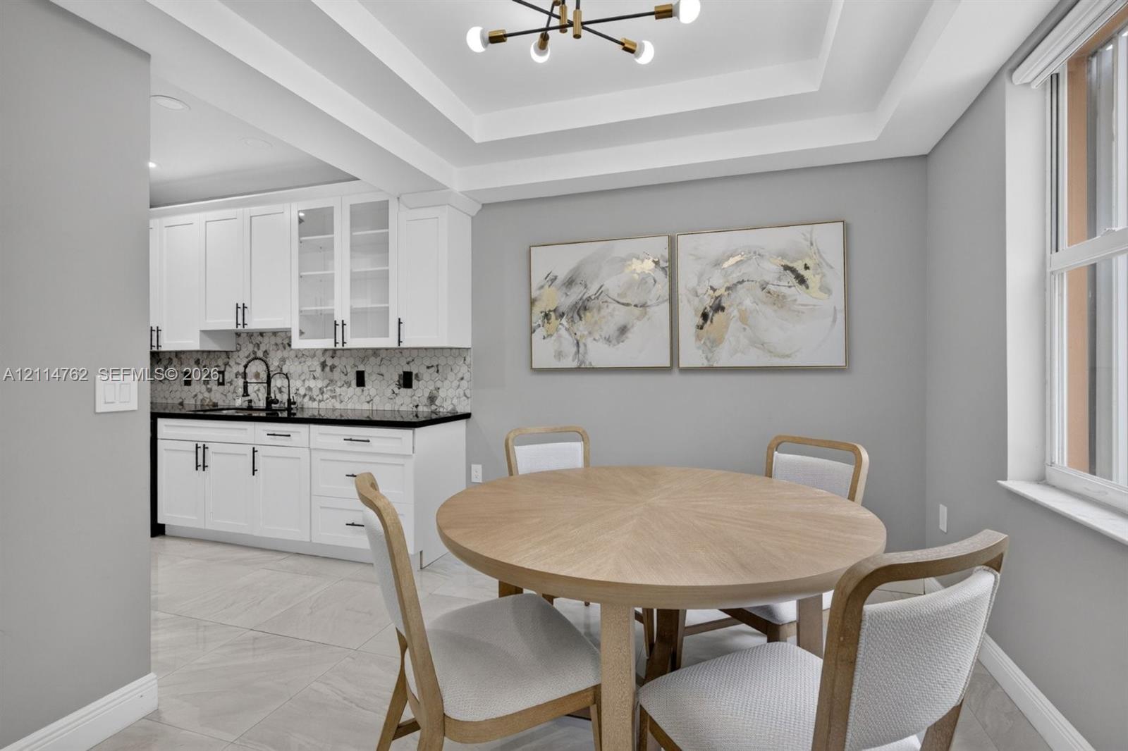 Tray ceiling with integrated lighting, white shaker cabinetry with glass-front uppers, dark stone countertops, patterned tile backsplash, and large-format tile flooring