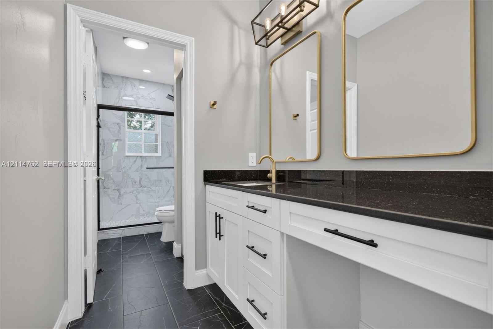 White shaker cabinetry with matte black hardware, dark countertop, dual mirrors with gold-tone frames, and large-format tile flooring