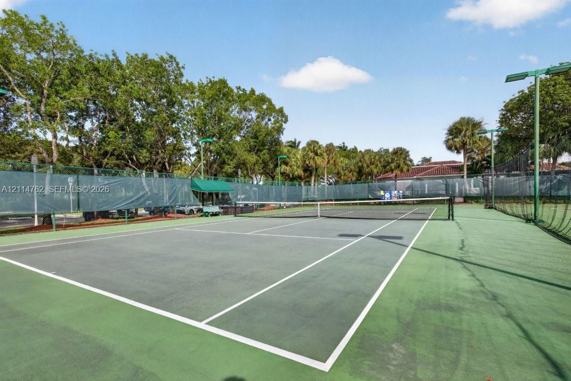 Hard-surface tennis court featuring perimeter chain-link fencing, protective mesh wind screening, integrated stadium-style lighting, and surrounding mature trees
