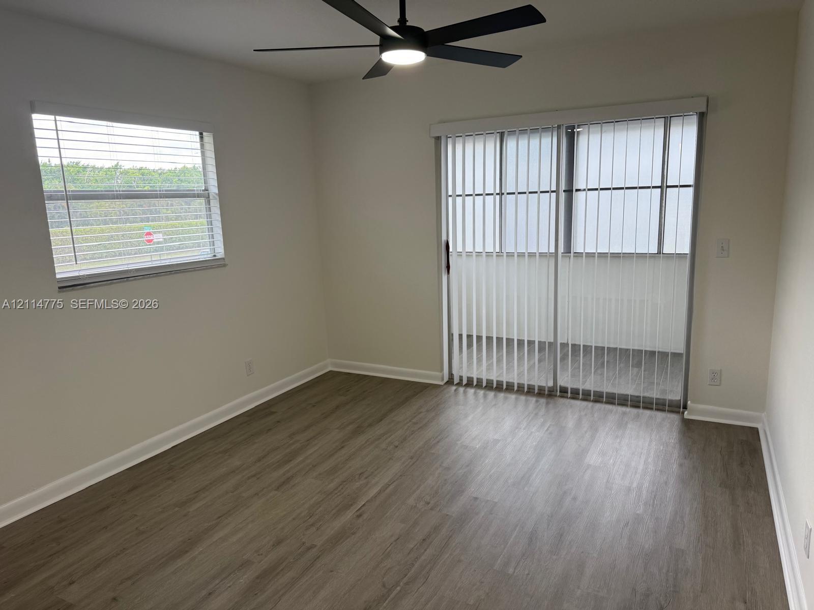 Room featuring wood-finish flooring, a matte black ceiling fan with integrated lighting, horizontal window blinds, and sliding glass doors with vertical blinds