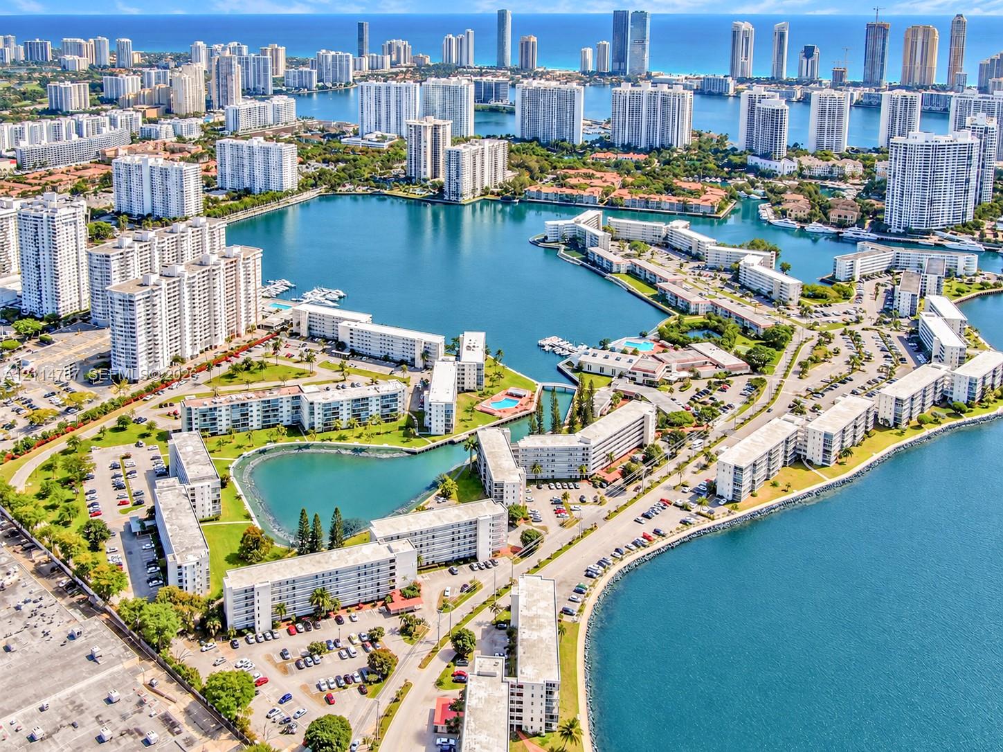 Aerial perspective of a multi-building residential community featuring direct waterfront access, paved surface parking areas, manicured green spaces, and a landscaped central lagoon