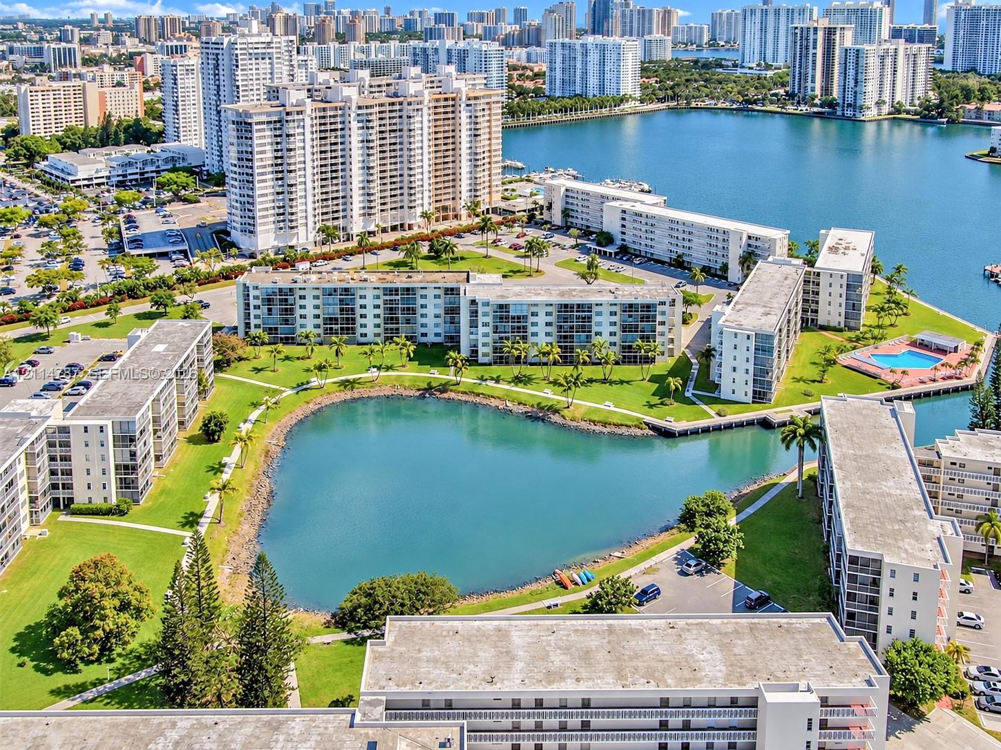 Aerial perspective of a multi-building residential complex featuring a central lake, landscaped grounds with mature palm trees, paved pathways, and an outdoor swimming pool