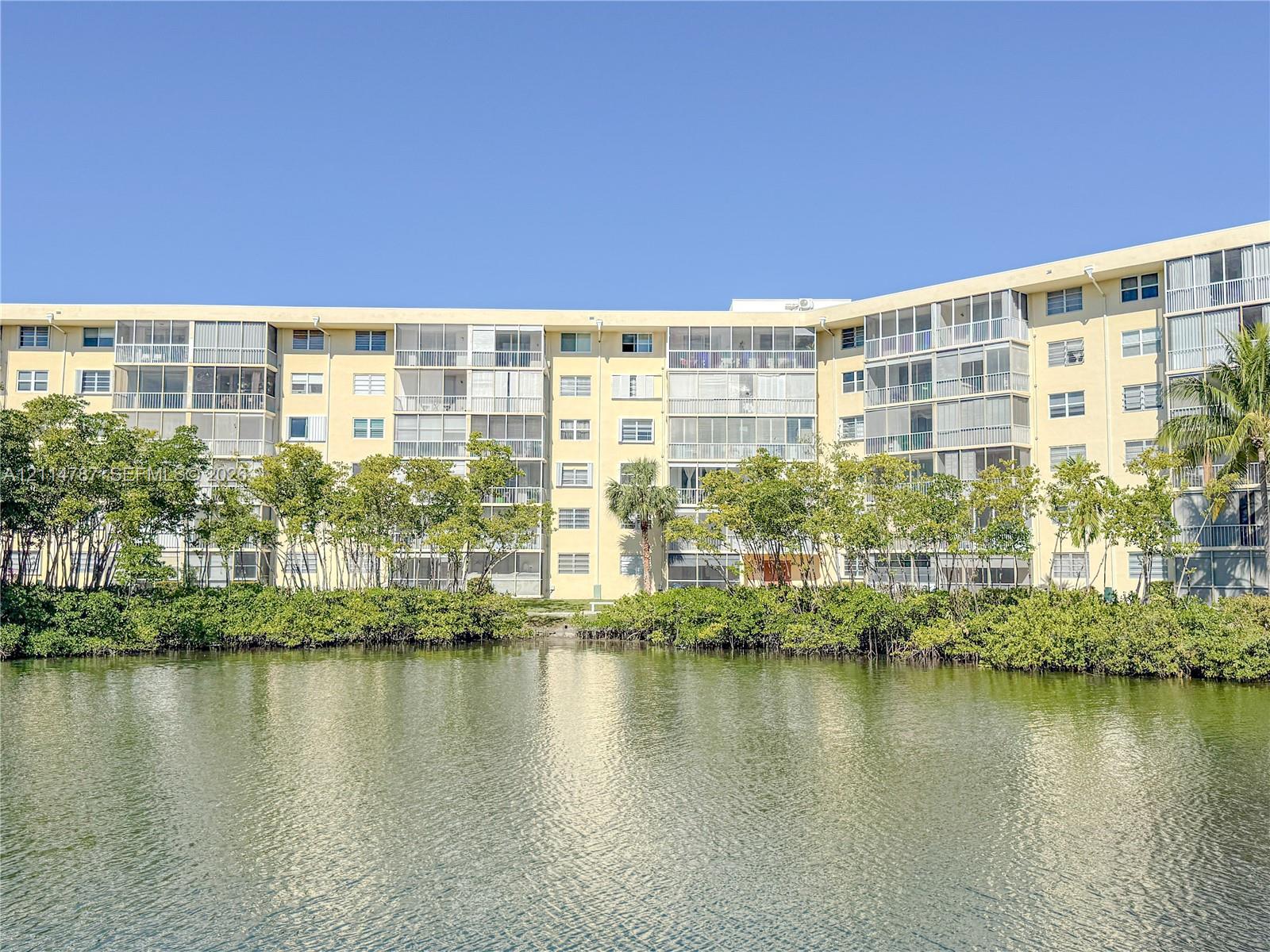 Multi-story residential building featuring screened balconies, stucco exterior, and waterfront placement with mature trees