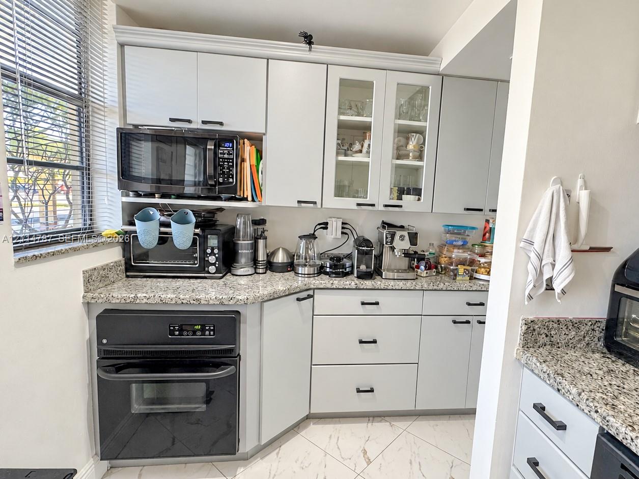Light gray shaker-style cabinetry with glass-paneled uppers, stone-patterned countertops, integrated wall oven, and tile flooring