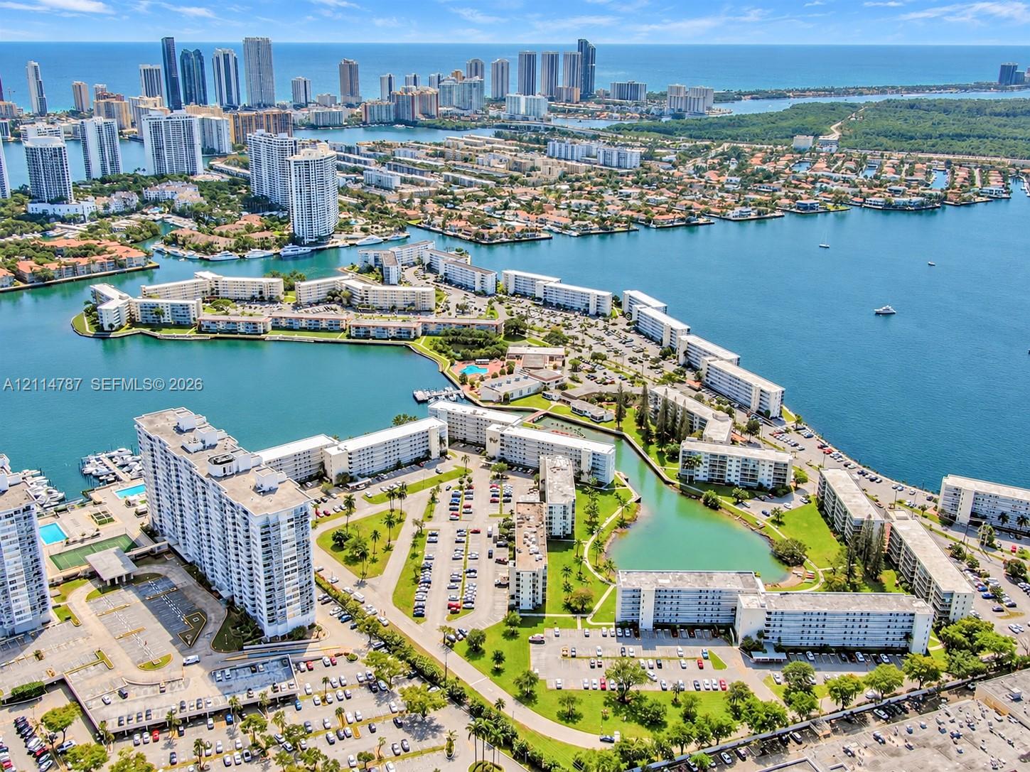 Aerial perspective of a waterfront residential complex featuring multiple mid-rise buildings, manicured grounds, peripheral parking areas, and expansive water frontage