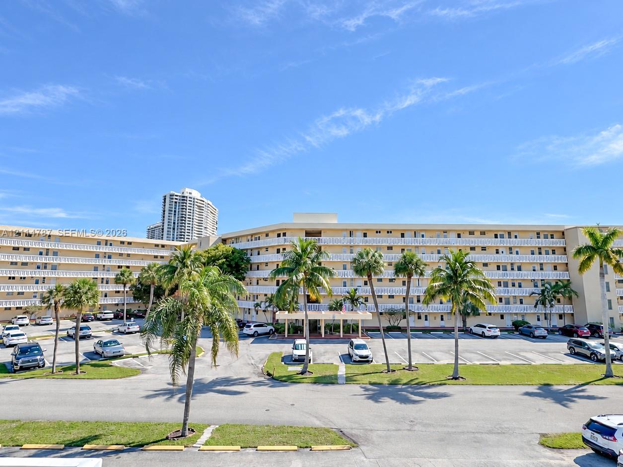 Multi-story residential complex featuring continuous exterior balcony railings, a central covered porte-cochre entry, surface parking, and mature palm tree landscaping