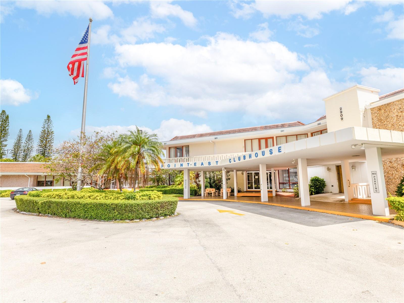 Clubhouse exterior featuring a covered porte-cochre, stone masonry facade, manicured landscaping, and asphalt parking area
