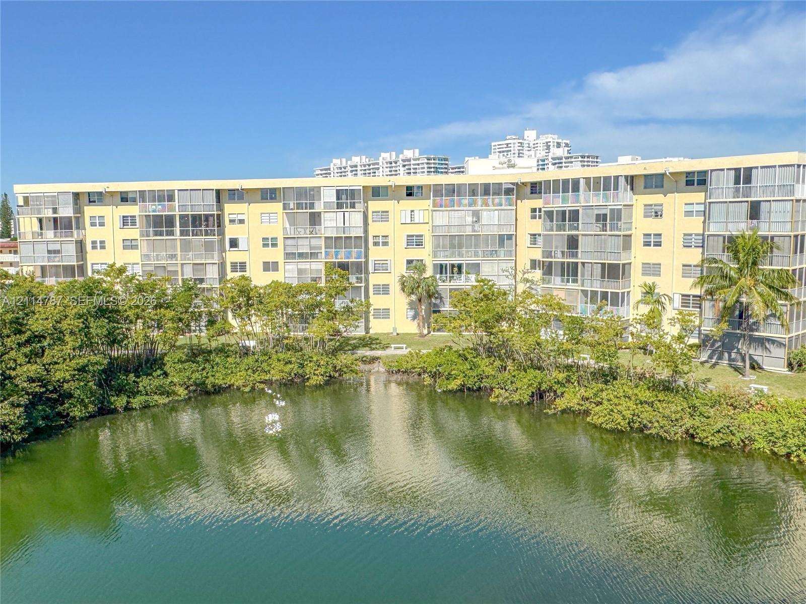 Multi-story building featuring screened balconies, light-colored exterior facade, mature landscaping, and waterfront positioning