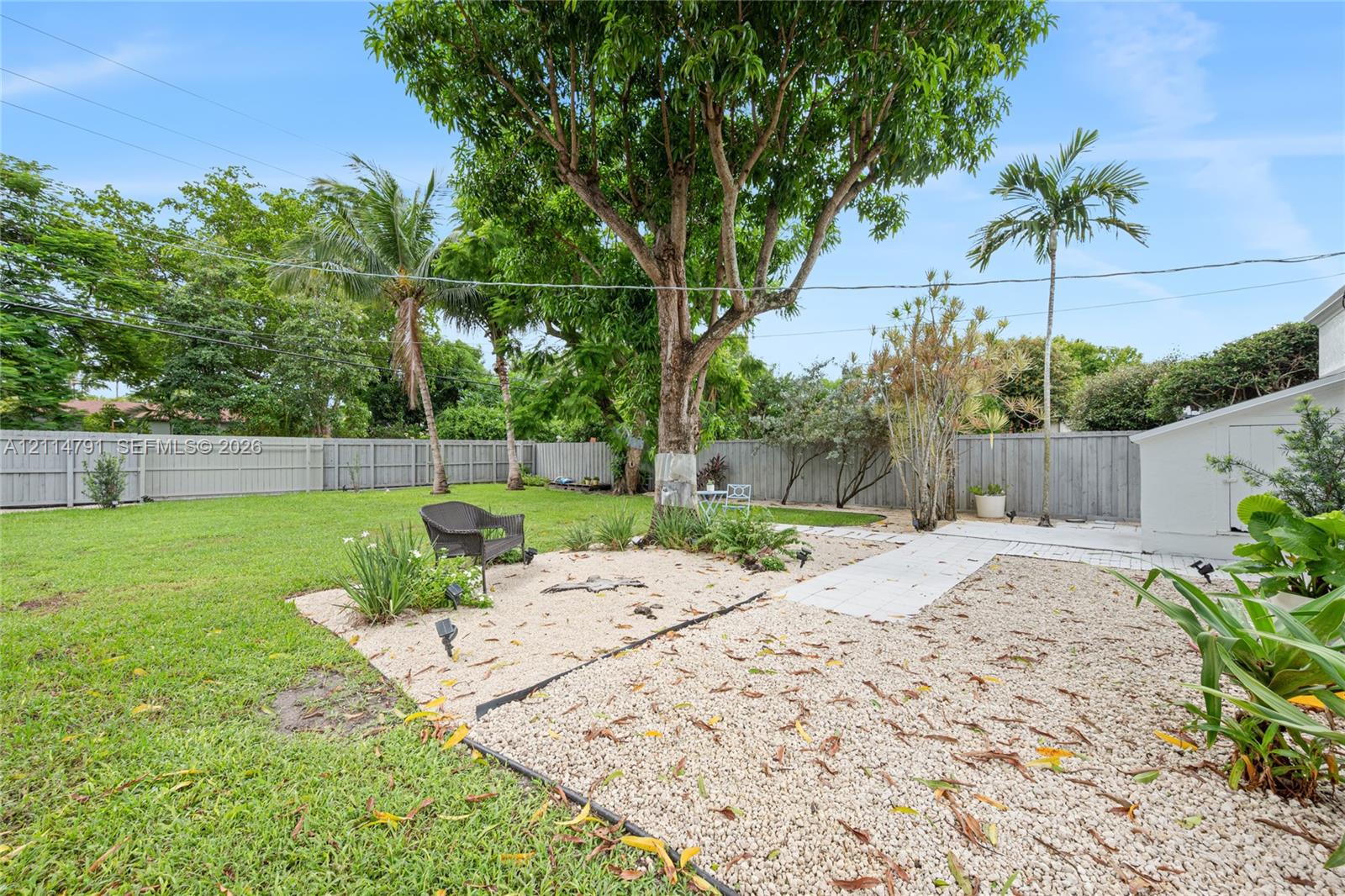 Fenced backyard featuring a manicured lawn, stone hardscaping, mature trees, and palm vegetation