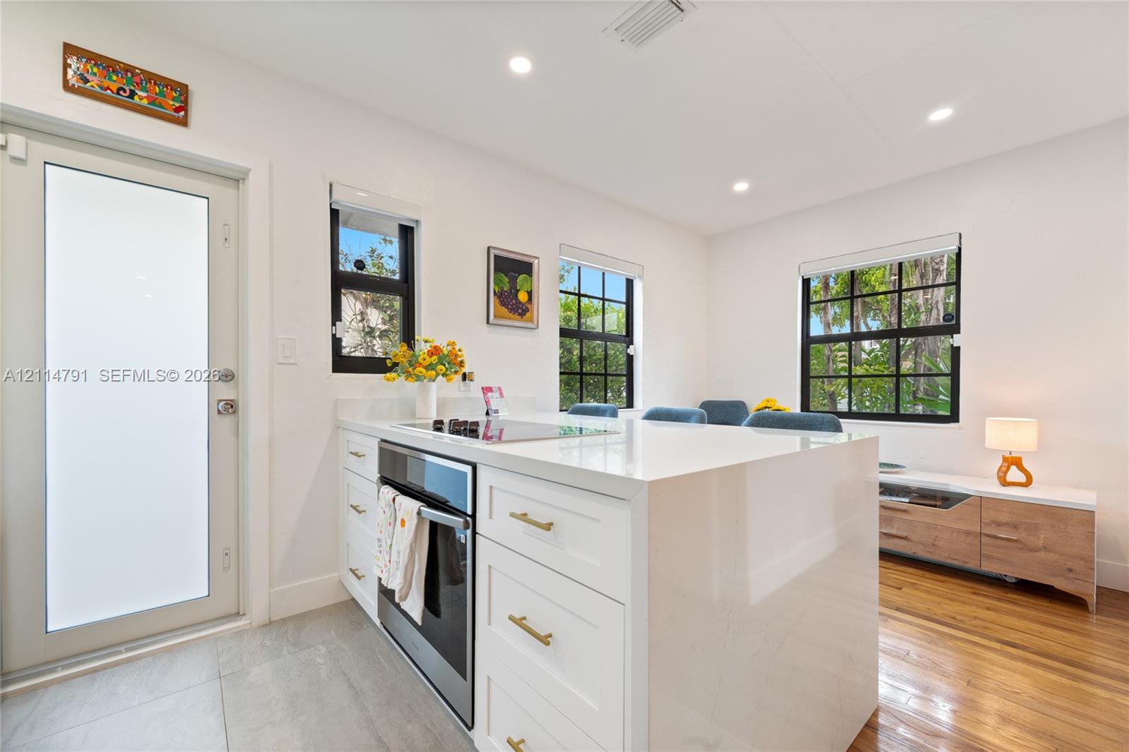 Kitchen featuring a waterfall-edge island with solid-surface countertops, shaker-style cabinetry with brass-tone hardware, built-in stainless steel oven, wood-finish flooring, and multi-pane windows with black frames