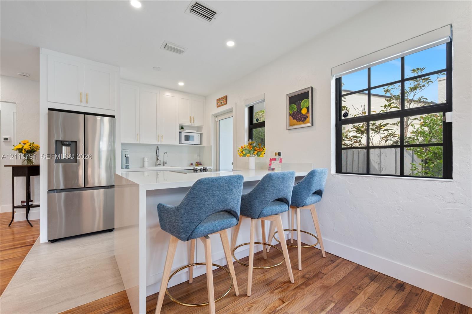 Kitchen featuring white shaker-style cabinetry, a waterfall-edge island with a stone-finish countertop, stainless steel refrigerator, recessed lighting, and a multi-pane window with a black frame