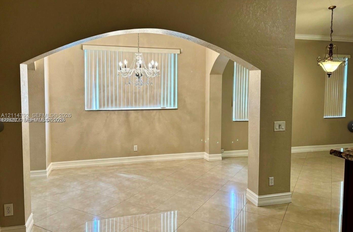 Interior space featuring prominent arched architectural openings, large-format tile flooring, dual multi-tiered crystal chandeliers, and multiple windows with vertical blinds